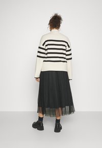 Mustavalkoraidallinen neulepaita, jossa on korkea kaulus, yhdistettynä liehuvaan mustaan laskoshelmaiseen hameeseen. Varustettu paksupohjaisilla mustilla kengillä.