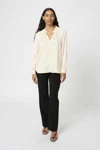 Lys beige langærmet bluse med V-hals og knaplukning, parret med skræddersyede sorte bukser og sorte remsandaler. Glat stofstruktur.