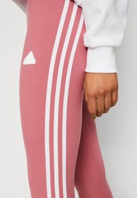 Leggings rosa claro com design de três riscas brancas, apresentando o logo da Adidas em branco na coxa. Tecido suave com um ajuste justo.