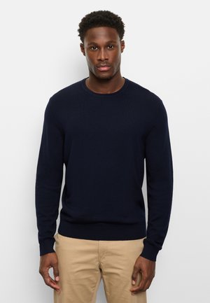 Man with short curly hair wearing a navy blue long-sleeve sweater and beige pants standing against a plain white background.