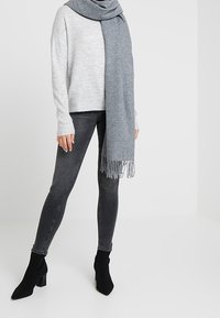 Grijze gebreide trui, donkergrijze spijkerbroek en een lichtgrijze sjaal met franjes. Zwarte enkellaarsjes maken de outfit af. Eenvoudige, casual look.