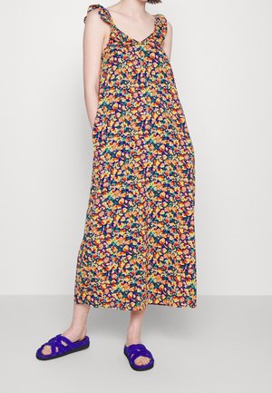Bloemenmaxi-jurk met ruches op de schouders, met een kleurrijk patroon van gele, oranje en blauwe bloemen op een donkere achtergrond. Gemaakt met paarse sandalen.