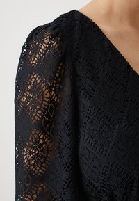 Μαύρη δαντελένια μπλούζα με περίπλοκα λουλουδάτα σχέδια, V-λαιμόκοψη και φουσκωτά μανίκια. Η υφή είναι λεπτή και διάφανα σχεδιασμένη.