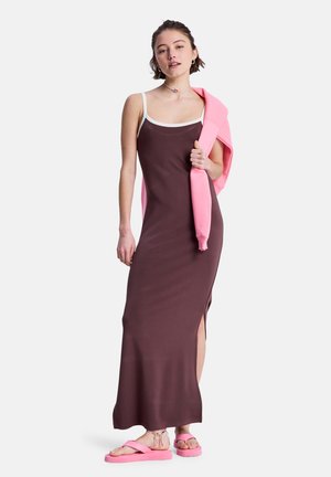 Braunes Maxikleid mit seitlichem Schlitz, dünnen Trägern und weißem Besatz. Kombiniert mit pinkfarbenen Sandalen und einem rosa Pullover, der über der Schulter getragen wird.