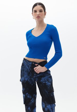 Blau gestreiftes Strickoberteil mit langen Ärmeln, V-Ausschnitt und cropped Design, kombiniert mit schwarzen und blauen Batik-Cargo-Hosen mit seitlichen Taschen.