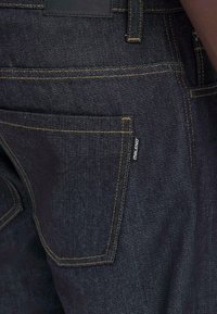 Jeans in denim blu scuro con cuciture gialle a contrasto, dotati di una tasca posteriore e di un piccolo etichetta vicino alla tasca. Texture liscia.