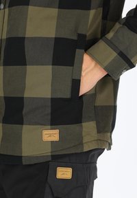 Green and black checked flannel shirt with a pocket, featuring a brown label. The fabric has a soft texture; buttons visible at the cuff.
