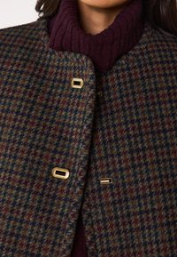 Cappotto in misto lana con un motivo a quadri scuri nei toni del verde, burgundy e blu. Chiuso con un hardware rettangolare dorato. Sotto, un dolcevita burgundy a costine.