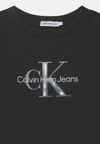 Musta collegepaita, joka on valmistettu pehmeästä kankaasta. Etuosassa on hohtava hopeinen logo, jossa lukee "Calvin Klein Jeans" ja "CK".