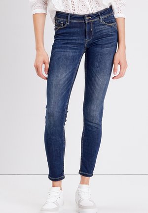 Jeans skinny en denim bleu foncé, avec une taille mi-haute, des poches avant et un léger délavage. Associés à des baskets blanches.