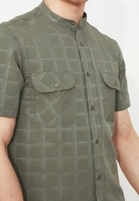 Vihreä lyhythihainen paita, jossa on kaulusta vailla oleva bandikaulus, ruutukuosisena, kahdella etutaskulla ja nappikiinnityksellä. Materiaali vaikuttaa kevyeltä.