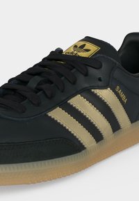 Μαύρο sneaker Adidas Samba με χρυσές ρίγες, χρυσό λογότυπο στη γλώσσα, κείμενο «SAMBA» στο πλάι, και σόλα από καουτσούκ.