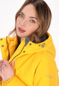 Gelbe wasserdichte Jacke mit einer grauen Fleece-fütterten Kapuze, ausgestattet mit einem Reißverschluss, Druckknöpfen und einem Logo auf dem Ärmel. Glatte Textur.
