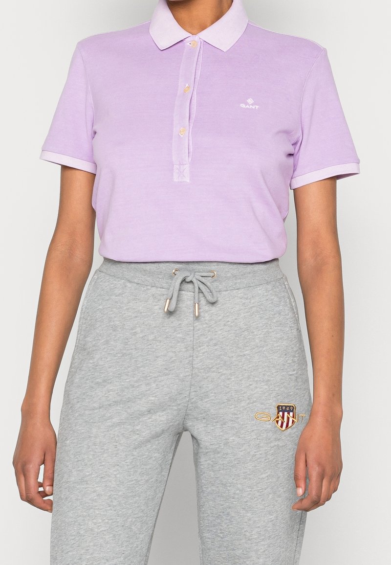 Polo shirt violet clair avec col et quatre boutons ; associé à un pantalon de survêtement gris avec un logo brodé et des accents dorés.