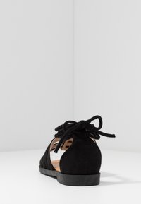 Sandale plate en suède noir avec lien à nouer à l'arrière, côtés ouverts et semelle en caoutchouc plate. Design minimaliste avec une forme structuré.