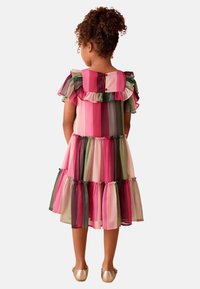 Ragazza in piedi, di spalle, che indossa un vestito a righe colorate con volant e maniche corte, abbinato a ballerine dorate su uno sfondo neutro.