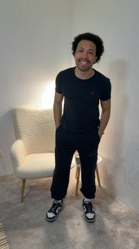 Mand med krøllet hår smiler, iført sort t-shirt og bukser, står i et tæppet rum med en hvid lænestol og et lille rundt bord med en kop.