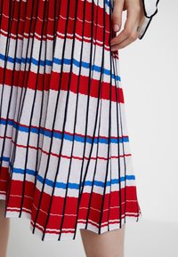 Gebreide jurk met horizontale strepen in rood, wit en blauw. Voorzien van plooien, dunne zwarte lijnen en een lichte uitlopende zoom.