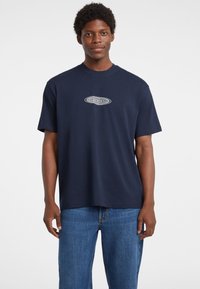 Granatowa bawełniana koszulka z krótkim rękawem, z logo "GUESS JEANS" w białym owalnym wzorze na klatce piersiowej.