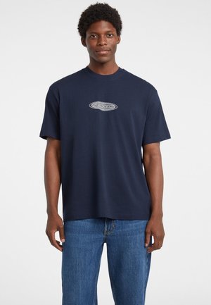 Granatowa bawełniana koszulka z krótkim rękawem, z logo "GUESS JEANS" w białym owalnym wzorze na klatce piersiowej.