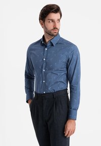 Camisa de manga larga azul con un sutil patrón tropical, que presenta un cuello abotonado y botones blancos. Combinada con pantalones oscuros de textura.