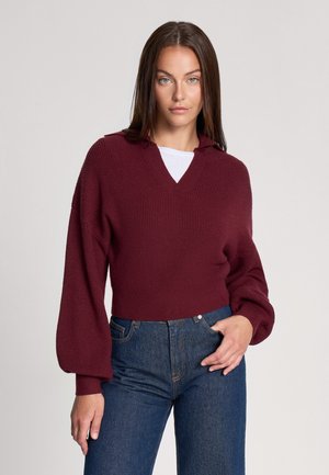 Maglione corto bordeaux con scollo a V e maniche a campana, texture a coste, abbinato a jeans scuri a vita alta.