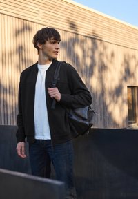 Svart bomberjacka med dragkedja över en vit t-shirt, bärs med mörka jeans. En svart väska hänger över ena axeln med detaljer i dragkedja och nät.