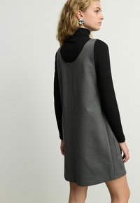 Vestido gris sin mangas en un material suave y texturizado, con escote redondeado y bolsillos laterales, llevado sobre una parte superior negra de manga larga.
