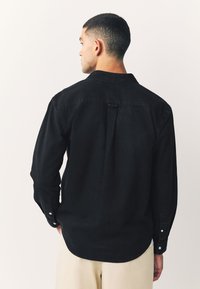 Schwarzes Langarmhemd mit einem Hemdkragen mit Knopfleiste, Rückendetail mit Falte und geknöpften Manschetten. Abgerundeter Saum, aus Baumwollstoff gefertigt.