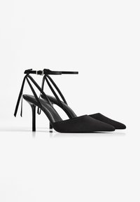 Scarpe nere con tacco alto dal design a punta, realizzate in materiale liscio, dotate di cinturini alla caviglia con fiocchi e un elegante tacco a stiletto.
