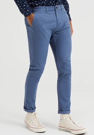 Mand iført slim-fit blå bukser rullet op ved anklerne og hvide høje sneakers, hænderne i lommerne, mod en ensartet lys baggrund.