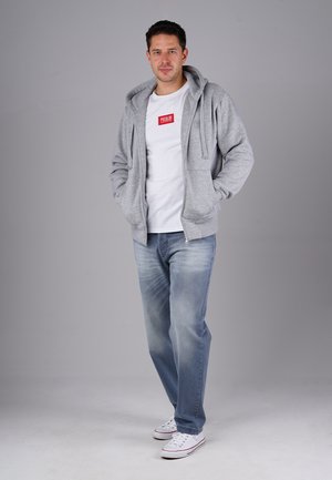 Ein Mann steht mit den Händen in den Taschen, trägt einen grauen Hoodie, ein weißes T-Shirt mit rotem Logo, blaue Jeans und weiße Sneakers vor einem einfarbigen grauen Hintergrund.