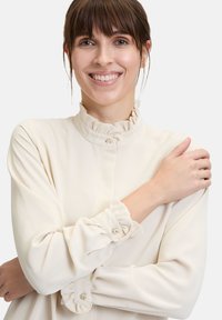 Crèmekleurige blouse met gerimpelde kraag en manchetten, met kleine ronde knopen. Glad textuur, aansluitend ontwerp, lange mouwen.