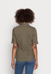 Olijfgroen shirt met korte mouwen en kraag, met een rugpand, subtiele textuur en splitten bij de mouwboorden. Draagt bij blauwe jeans.