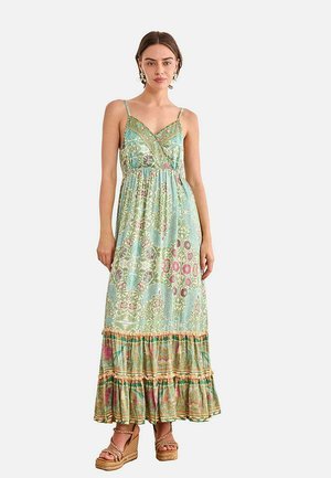 Mujer de pie vistiendo un vestido maxi sin mangas con estampado, bajo con volantes y sandalias de cuña con tiras en el tobillo, sobre un fondo liso.