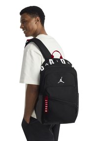 Mochila negra con un gran logo blanco de "JORDAN", etiqueta de cremallera roja y un logo Jumpman blanco. Cuenta con correas acolchadas y múltiples compartimentos.
