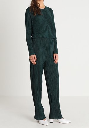 Grøn plisseret jumpsuit med lange ærmer og en samlet talje, der har lodret tekstur; båret med hvide spidse sko.