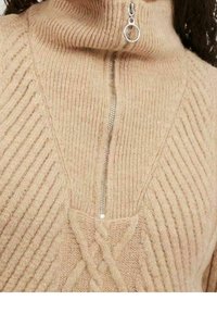 Maglione beige lavorato a maglia con colletto alto, caratterizzato da una cerniera argentata e una texture rigata verticale, arricchito da un motivo a treccia sul davanti.