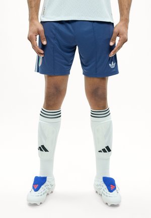 Osoba nosząca niebieskie sportowe szorty Adidas z odznaką Włoch, białe skarpetki Adidas do kolan z ochraniaczami na golenie oraz białe korki z niebieskimi nakładkami.