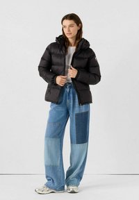 Giacca a vento nera con cappuccio, chiusura con zip e polsini elastici; abbinata a jeans a palazzo blu con patchwork e sneakers argento.