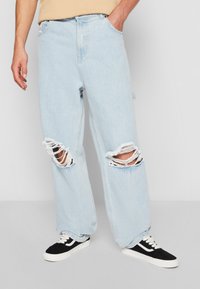 Pantalones vaqueros oversize color azul claro con rodillas rasgadas, mostrando bordes deshilachados. Combinados con zapatillas negras y blancas, que cuentan con un diseño clásico de corte bajo.