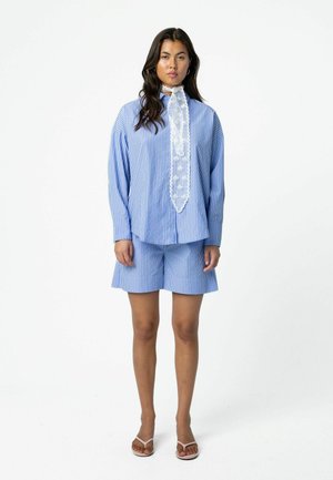 Vrouw staand, gekleed in een blauw-wit gestreept overhemd met lange mouwen en een witte kanten strik, bijpassende gestreepte korte broek en witte sandalen.