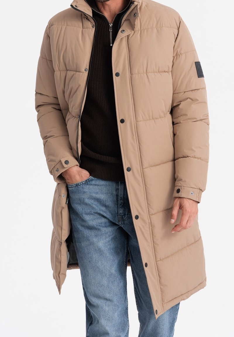 Manteau long matelassé beige avec boutons-pression, porté sur un pull noir à fermeture éclair, accompagné d'un jean bleu clair, main dans la poche, sur fond blanc uni.