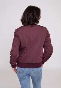 Giubbotto bomber bordeaux con orlo e polsini a costine, caratterizzato da una texture liscia e lucida e tasche laterali, indossato sopra jeans di denim blu.