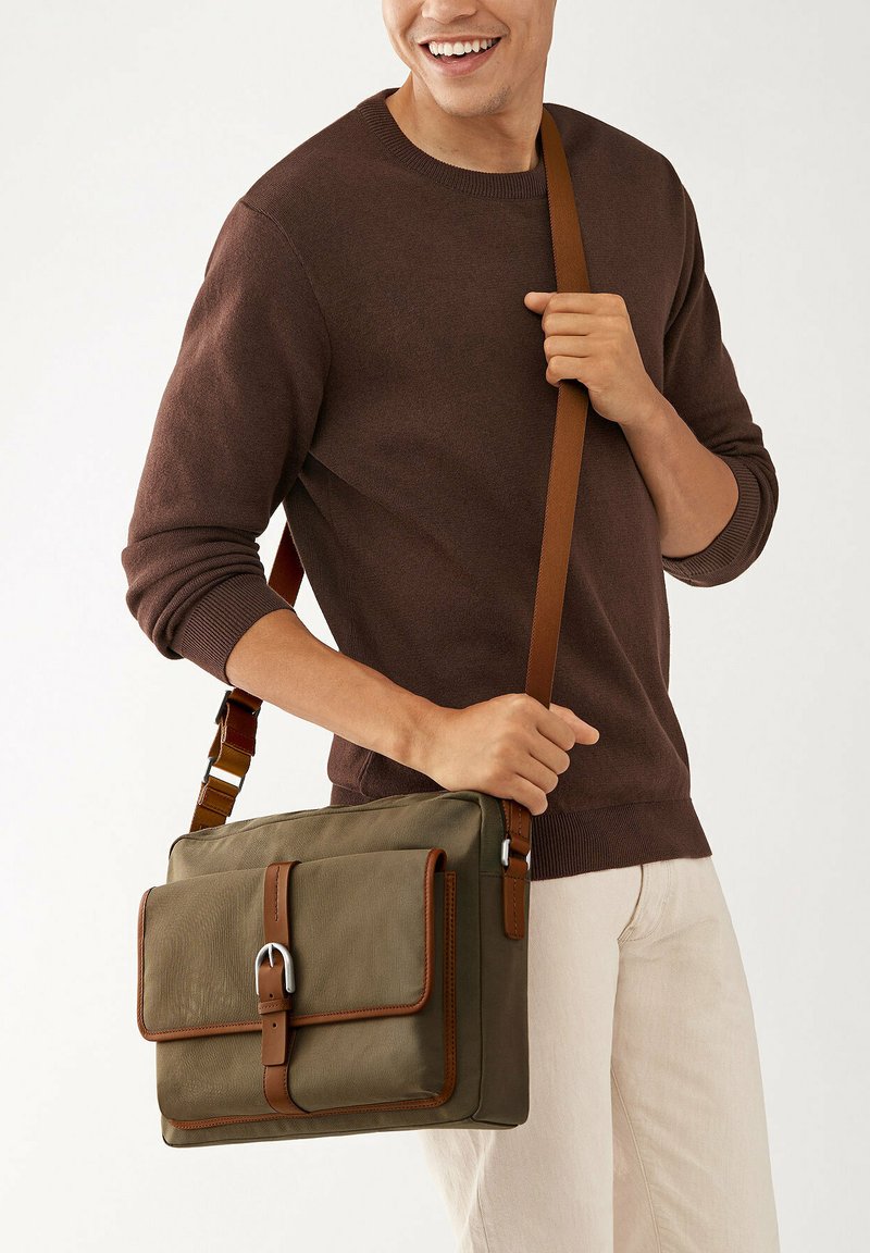 Olive green messenger bag with brown leather accents, rectangular shape, front buckle detail, and adjustable strap, held by a person.