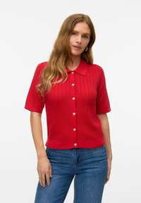 Röd stickad kortärmad kavaj med krage, med en vågig knappslå och ett strukturerat mönster genom hela plagget. Blå jeans kompletterar outfiten.