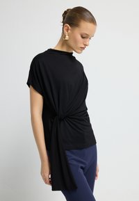 Mujer joven con el cabello recogido en un moño, vistiendo una blusa negra de manga corta anudada a la cintura y pantalones azules, de pie frente a un fondo blanco liso.