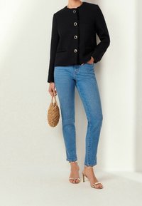 Vrouw in een zwart knoopjasje, blauwe skinny jeans, gouden bandjeshakken, met een kleine bruine geweven tas.