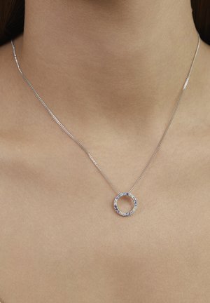 Silberne Halskette mit einem runden Anhänger, der blaue und klare Edelsteine enthält, an einer feinen Kette, präsentiert auf einem neutralen Hauthintergrund.