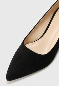 Scarpa nera in suede con tacco alto, punta affilata e scollatura bassa, dotata di una soletta beige liscia e un design semplice.